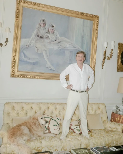 Indoor portrait of a man in white standing over a golden retriever on a tufted sofa beneath a large portrait painting.