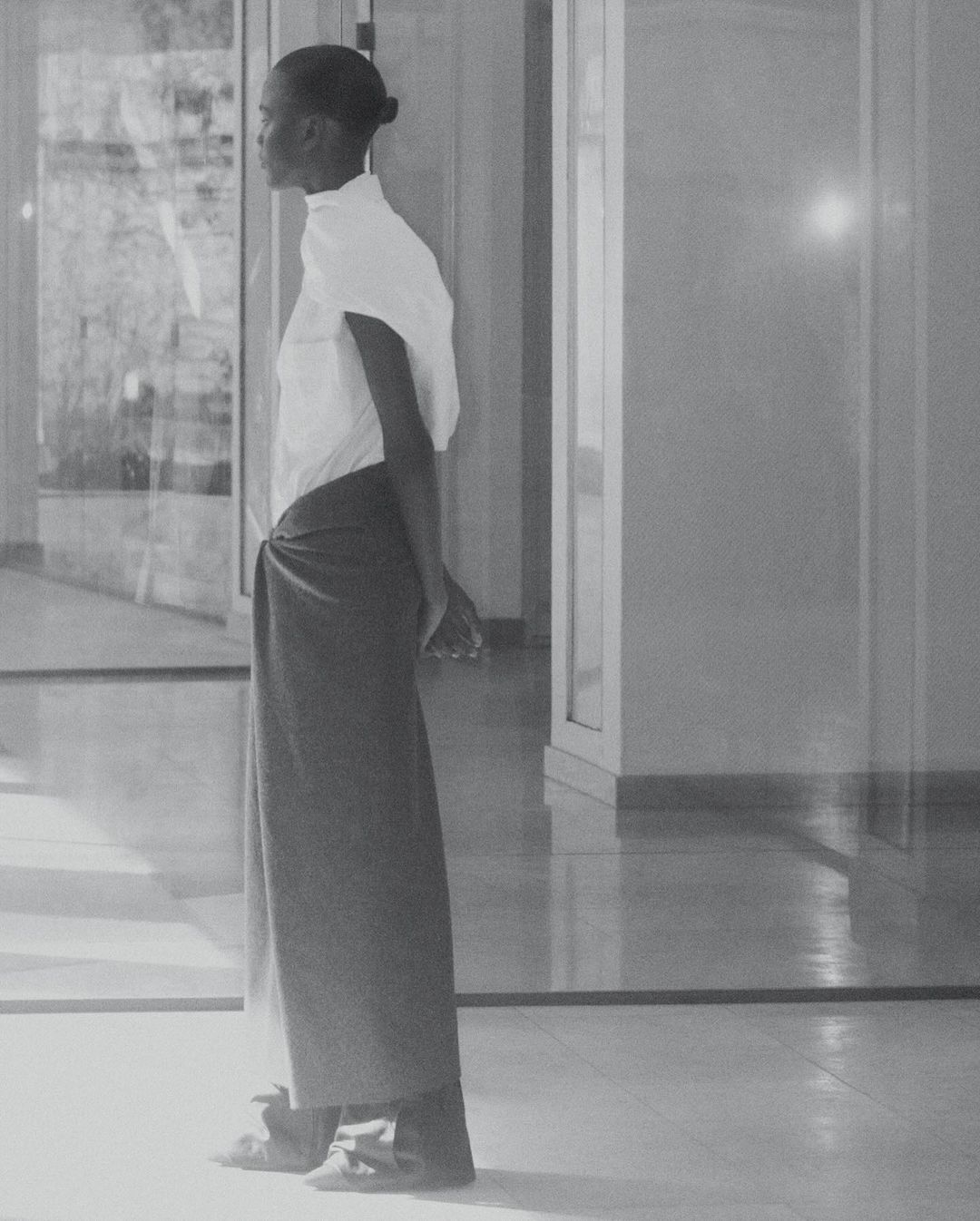 Woman in a white blouse and sarong stands in a bright, modern indoor space with glass doors, looking to the left in a side-view shot.