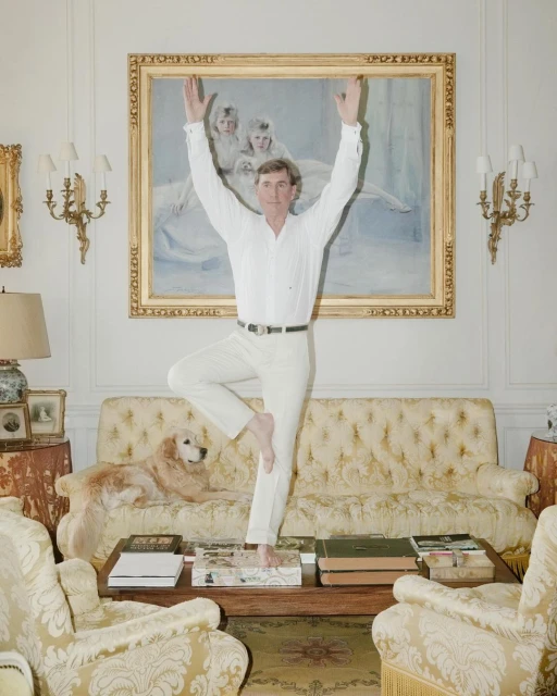 A man in white clothing practices a yoga pose indoors, balancing on one foot with arms raised, with a dog and traditional decor.