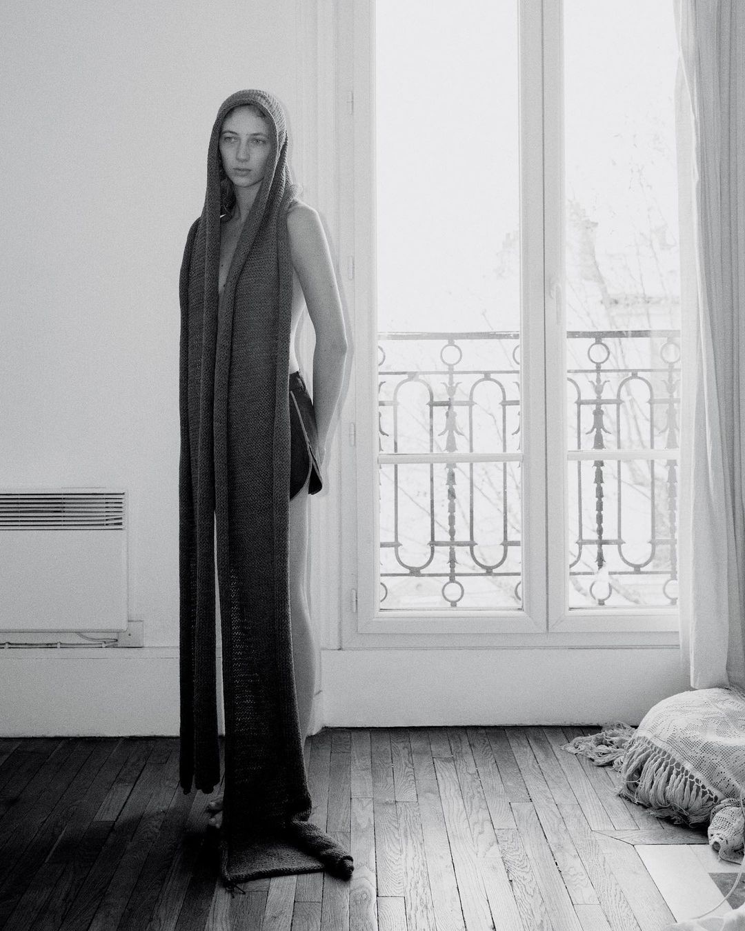 Black and white studio shot of a woman wearing a long knitted scarf with a hood, standing indoors by a window.