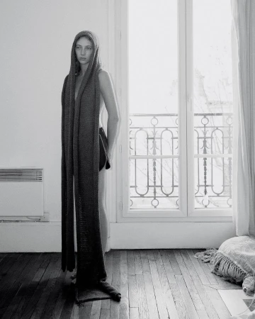 Black and white studio shot of a woman wearing a long knitted scarf with a hood, standing indoors by a window.