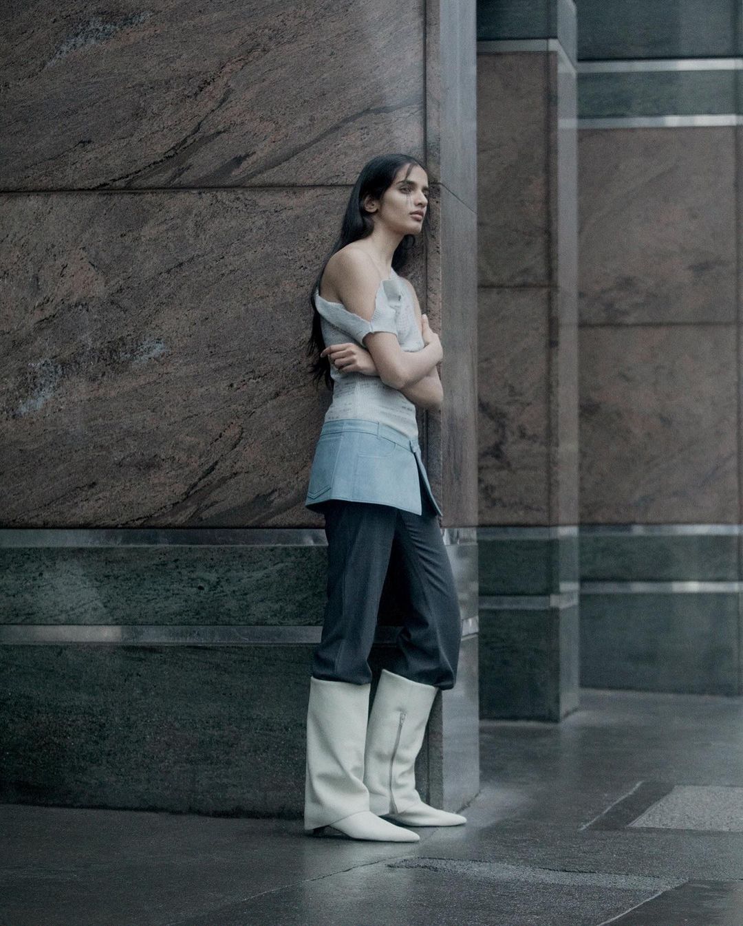 Indoor fashion portrait of an adult woman with long brown hair, wearing a light blue top, dark pants, and high white boots.