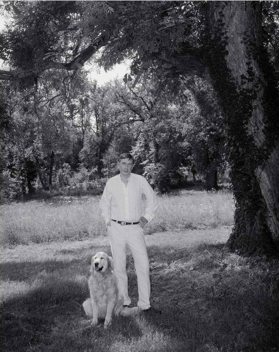Monochrome portrait of an adult man in a white outfit posing with a golden retriever outdoors in a grassy, wooded area with dappled light.