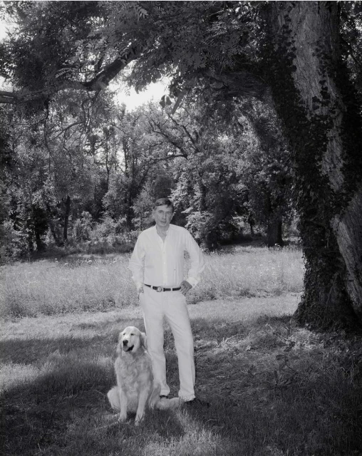 Monochrome portrait of an adult man in a white outfit posing with a golden retriever outdoors in a grassy, wooded area with dappled light.