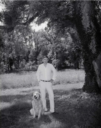 Monochrome portrait of an adult man in a white outfit posing with a golden retriever outdoors in a grassy, wooded area with dappled light.
