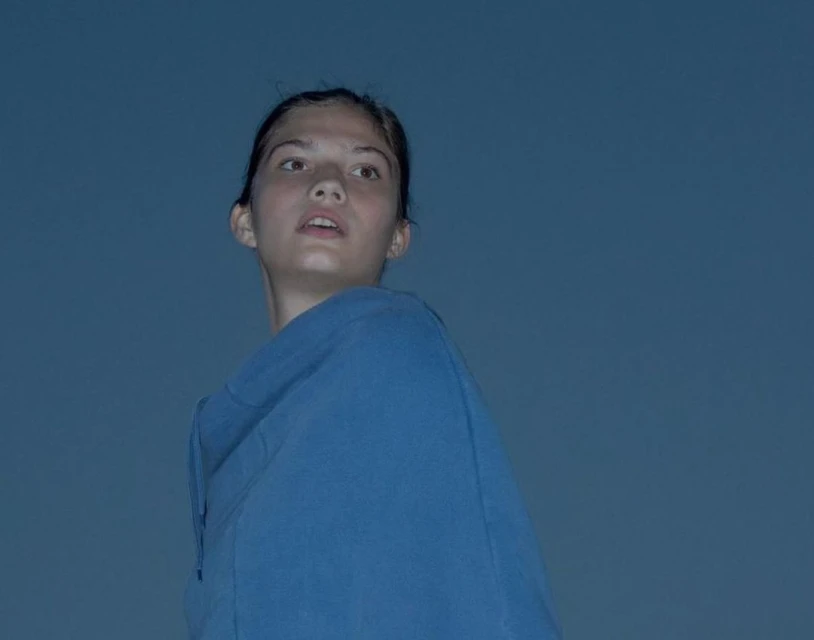 Young woman gazes upwards against a solid blue background, wearing a blue hoodie, in a low-angle portrait.