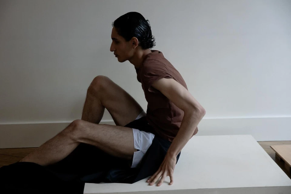 Adult male wearing a brown shirt, white underwear, and black bottoms sits indoors against a white wall on a white platform.