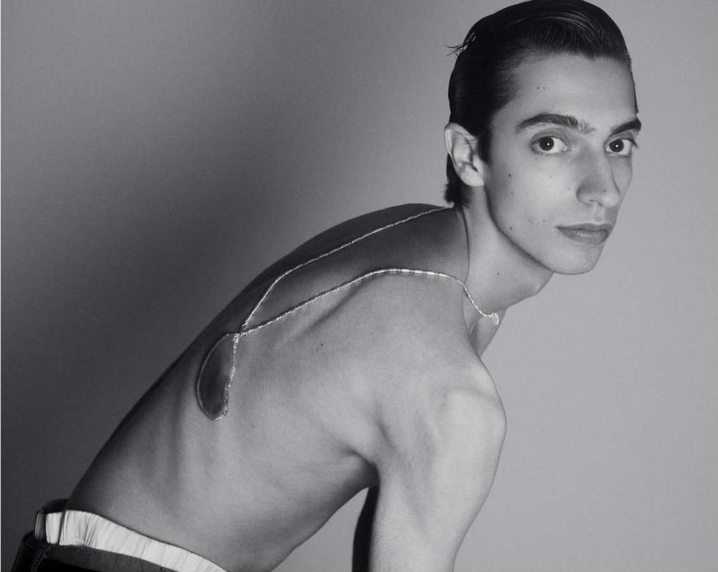 Monochrome portrait of a man wearing a metallic body jewelry piece draped across his bare back by Romain Duquesne