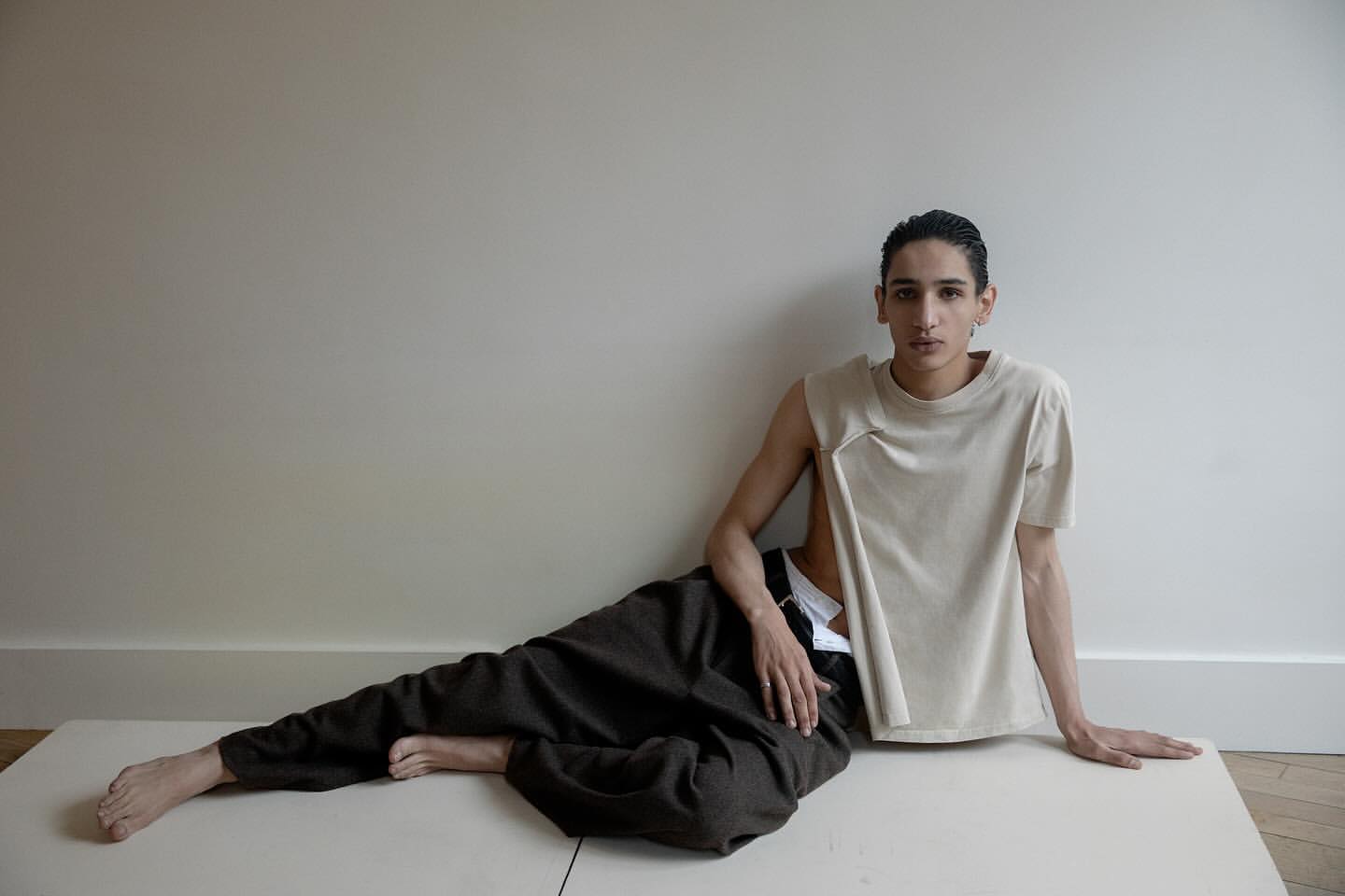 Person poses indoors wearing beige sleeveless top and dark pants, leaning against a white wall on a cream surface in a portrait shot.