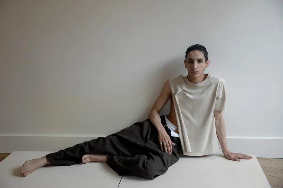 Person poses indoors wearing beige sleeveless top and dark pants, leaning against a white wall on a cream surface in a portrait shot.