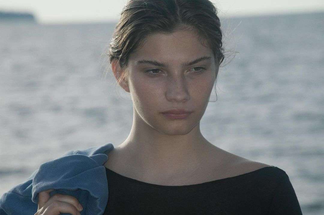 Young woman gazes downward against a backdrop of serene water, her hair pulled back and a denim jacket casually draped over her shoulder.