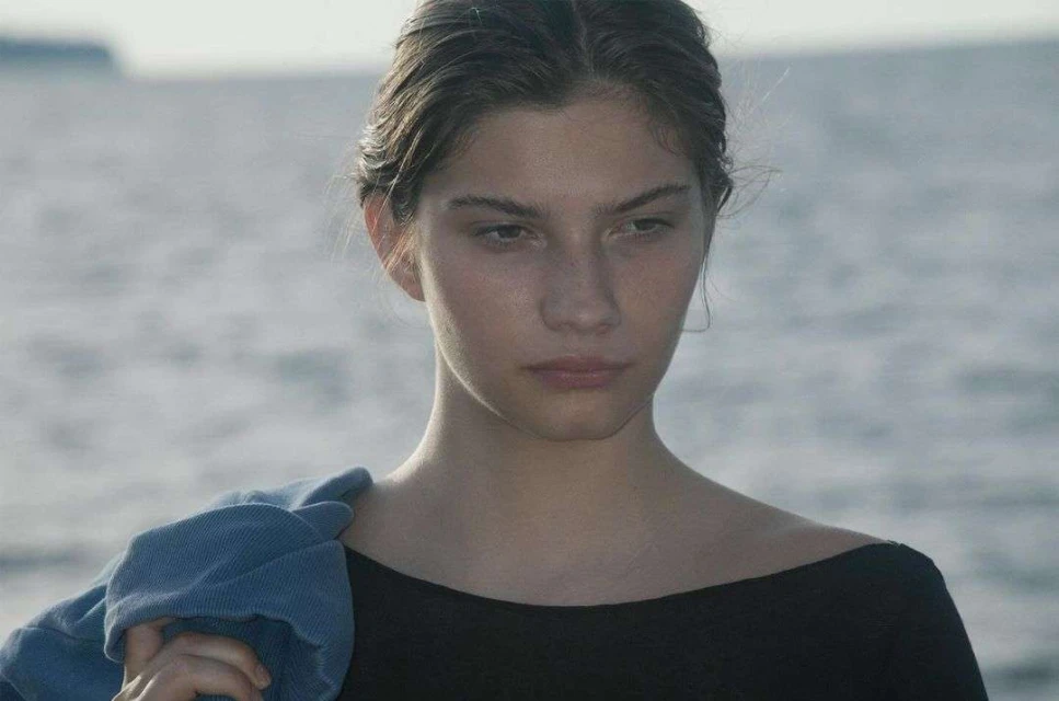 Young woman gazes downward against a backdrop of serene water, her hair pulled back and a denim jacket casually draped over her shoulder.