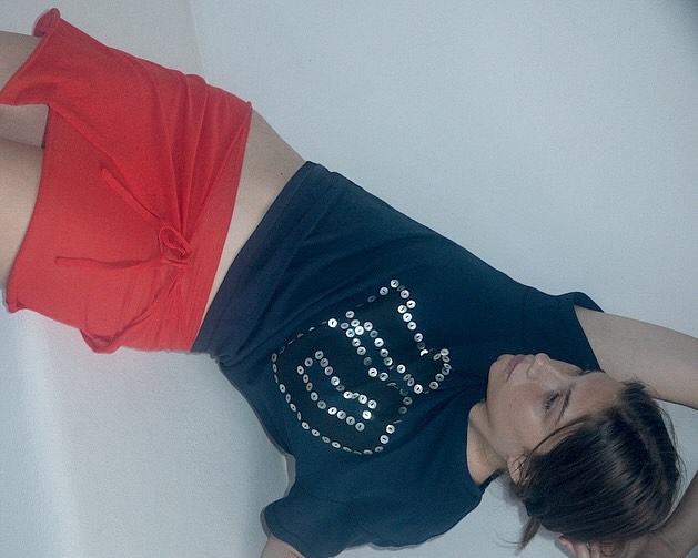 Woman poses in a black T-shirt with a sequin design and a red skirt indoors against a plain wall with a relaxed, casual style.