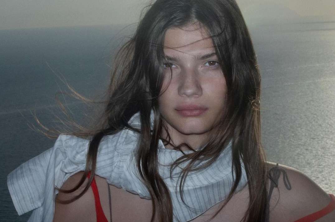 Close-up portrait of a woman with long brown hair wearing a white patterned scarf against an ocean background.
