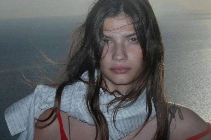 Close-up portrait of a woman with long brown hair wearing a white patterned scarf against an ocean background.