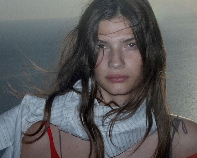 Portrait of a young adult woman with long brown hair in an open white shirt, gazing at the camera, against a background of ocean water.