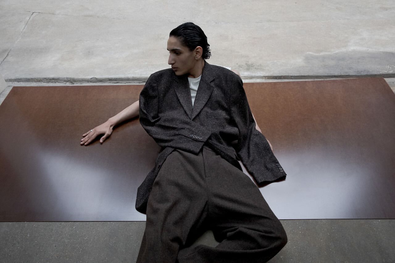 An adult male poses indoors wearing a brown jacket and pants, against a grey and brown background, in a fashion editorial style.