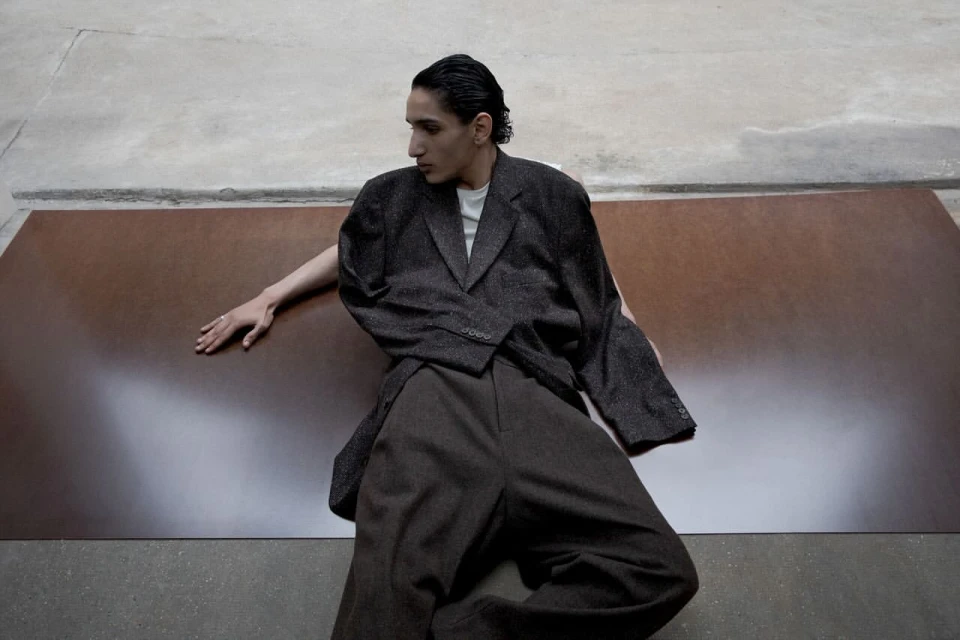 An adult male poses indoors wearing a brown jacket and pants, against a grey and brown background, in a fashion editorial style.