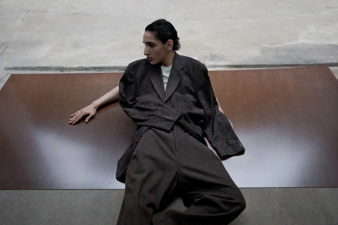 An adult male poses indoors wearing a brown jacket and pants, against a grey and brown background, in a fashion editorial style.
