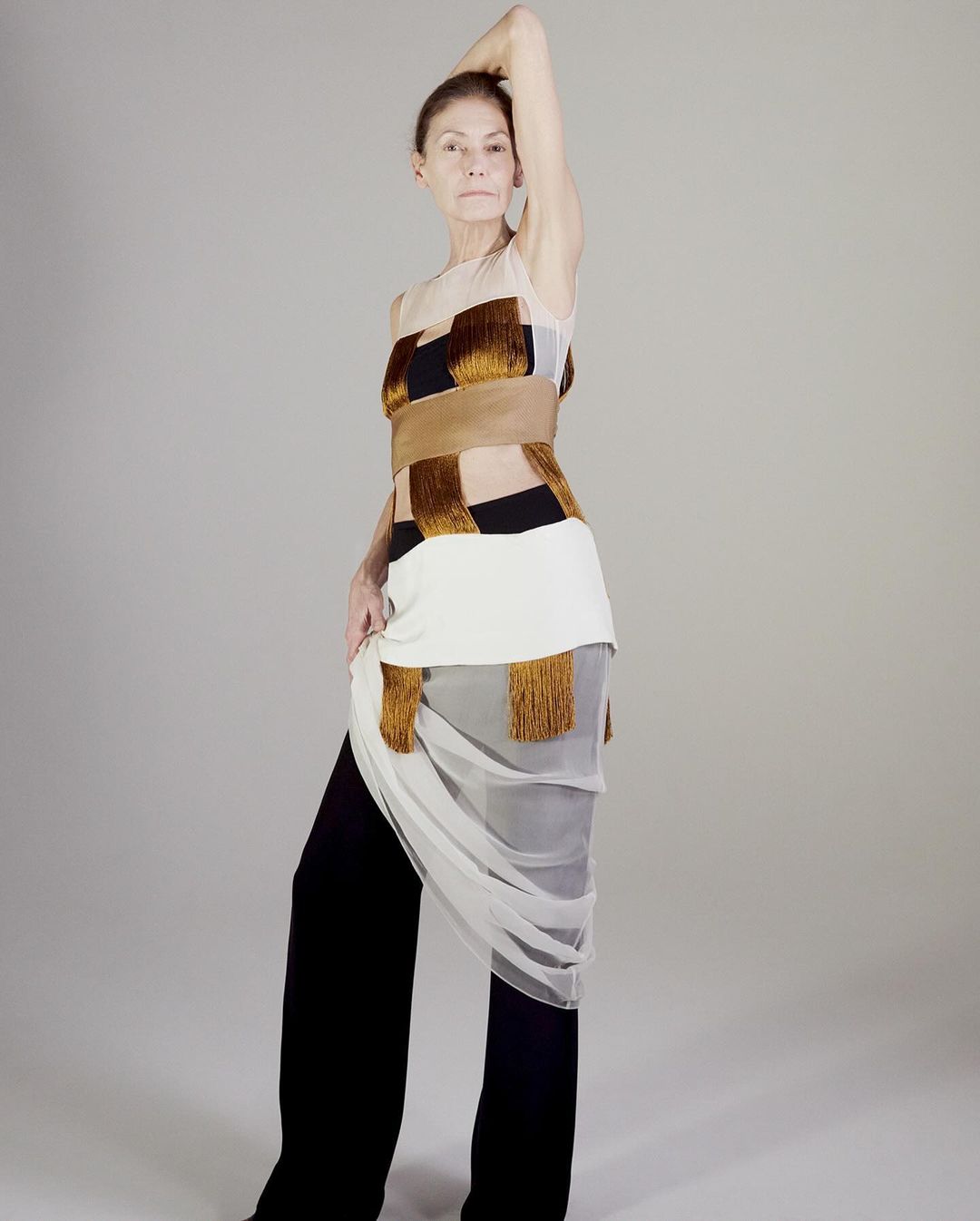Adult woman in a fashion pose wearing a light colored fringed top and black pants in a studio against a neutral background.