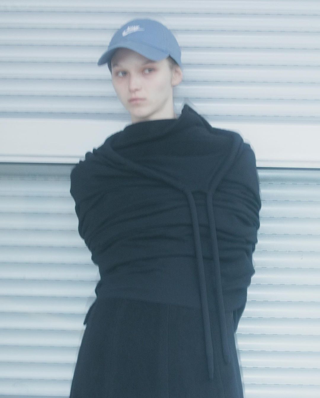 Woman in black dress and blue baseball cap poses against a corrugated metal background. Soft focus and minimalist fashion aesthetic.