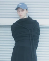 Model wearing a blue baseball cap and black wrapped top standing against a light grey shutter door in Paris.