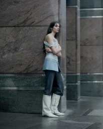 Woman leaning against a large stone pillar wearing an off-shoulder top, light blue wrap skirt, dark trousers, and white boots