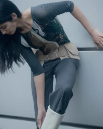 Model in grey top with graphic print, utility belt, and trousers leaning against a grey wall in Paris by Romain Duquesne