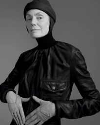 Monochrome portrait of an elderly woman wearing a black leather jacket, fitted head covering, and light-colored gloves against a gray background.