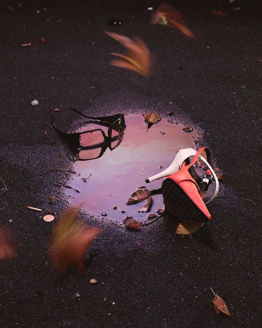 Sunglasses and a high-heeled shoe sit in a puddle on the ground, surrounded by fallen leaves, capturing a moment of urban disregard.