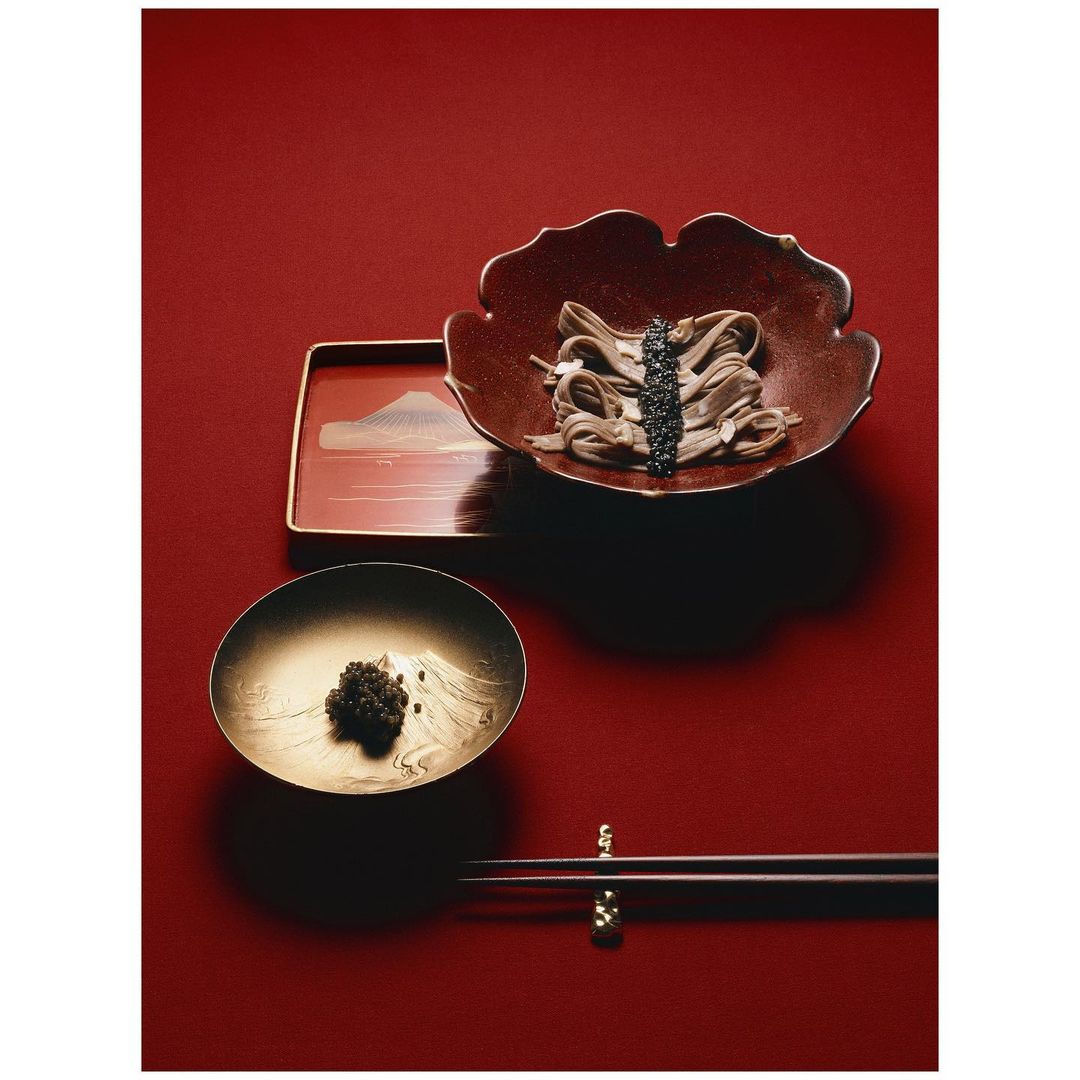 Overhead shot of soba noodles and caviar in decorative bowls with chopsticks against a deep red background, elegant food presentation.