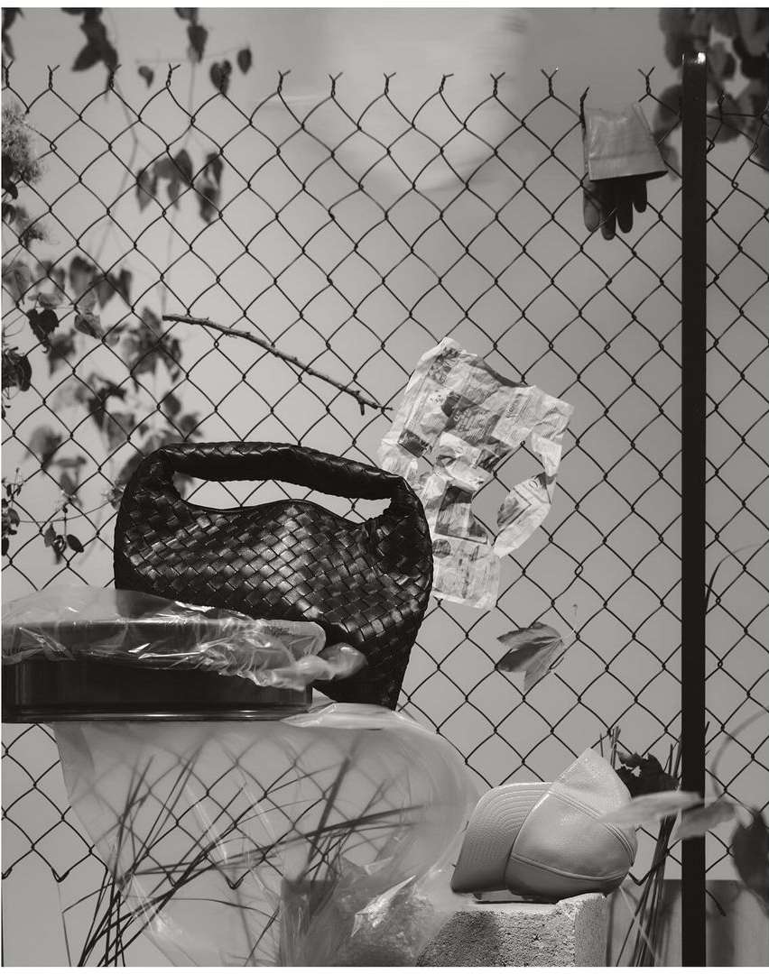 Still life with woven purse on a covered dish, plastic bags, and a hat on a concrete block against a chain link fence.