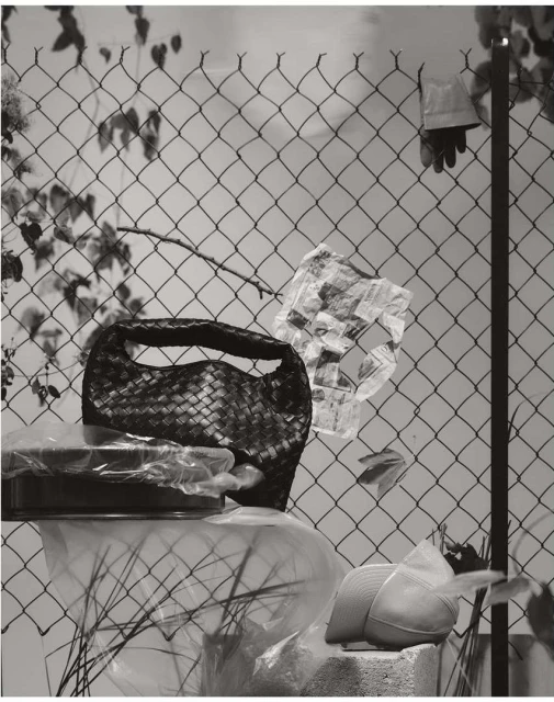 Still life with woven purse on a covered dish, plastic bags, and a hat on a concrete block against a chain link fence.