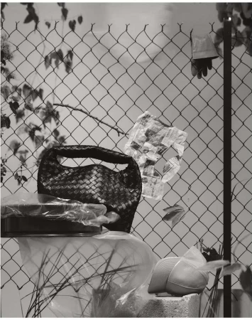 Still life with woven purse on a covered dish, plastic bags, and a hat on a concrete block against a chain link fence.