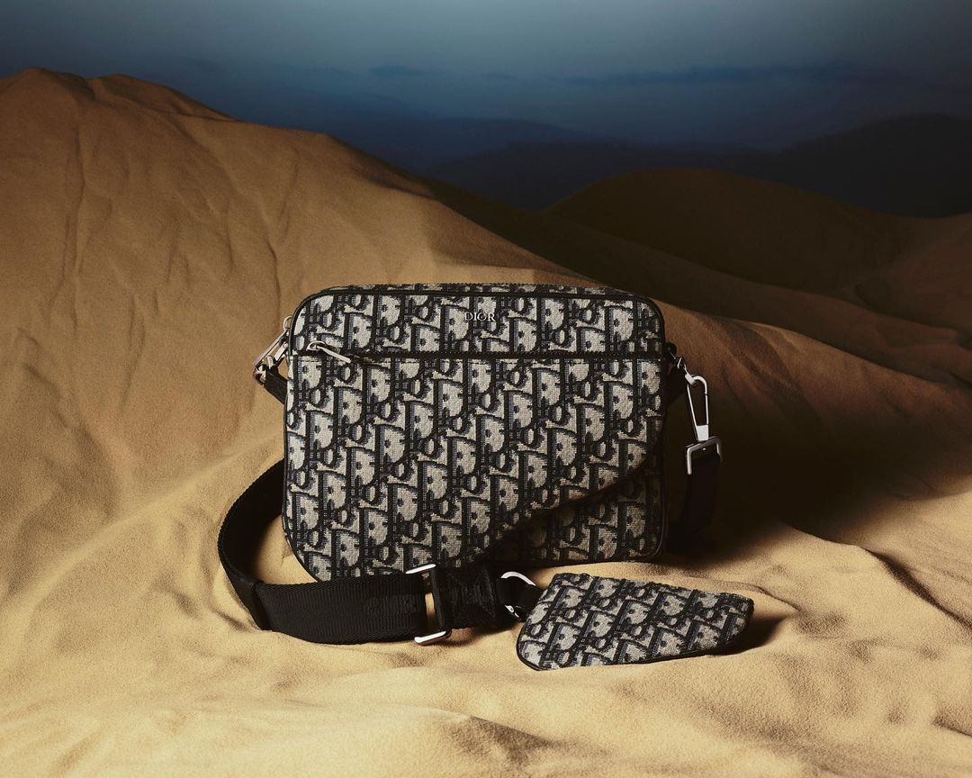 Luxury Dior Origin crossbody bag and pouch with allover logo print, laying on tan fabric against a landscape mountain background.