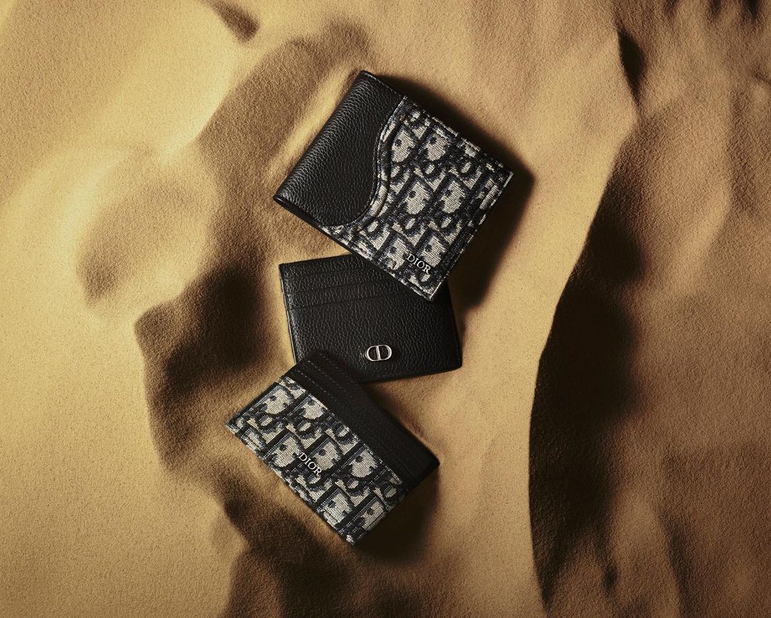 Three Dior wallets and cardholders with a patterned fabric and leather finish are displayed on a textured tan background.