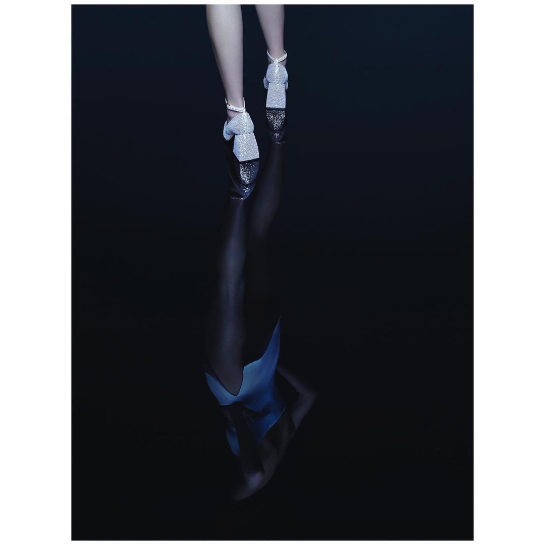 Legs of a person wearing sparkly high heels with ankle straps reflected on a glossy black surface in a dark fashion studio.