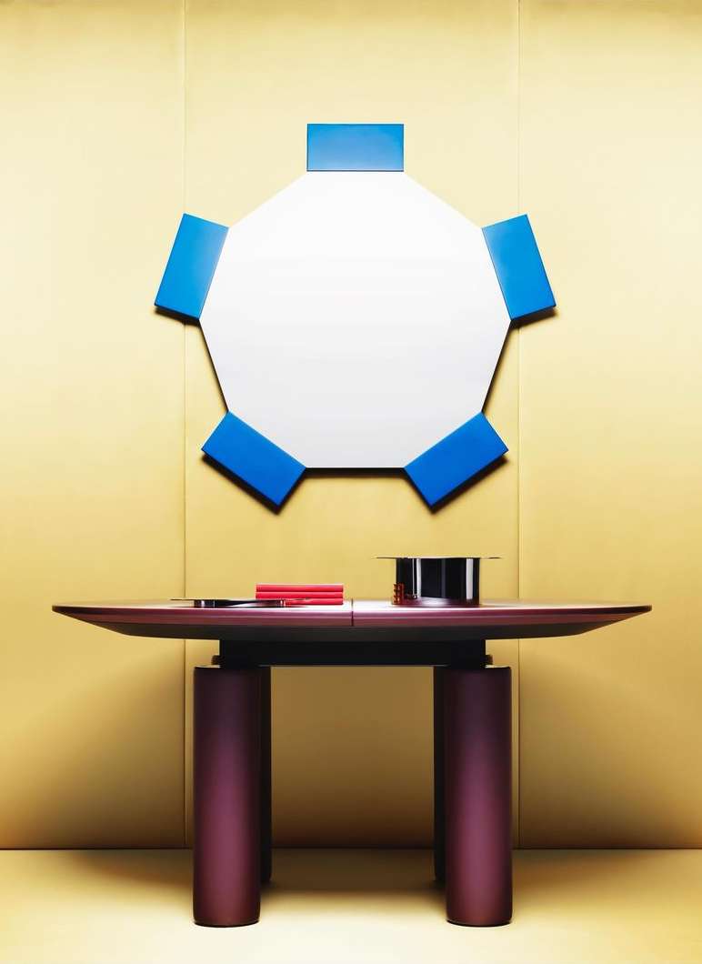Contemporary interior featuring a table with cylindrical legs, a geometric mirror with blue accents, and a gold background.
