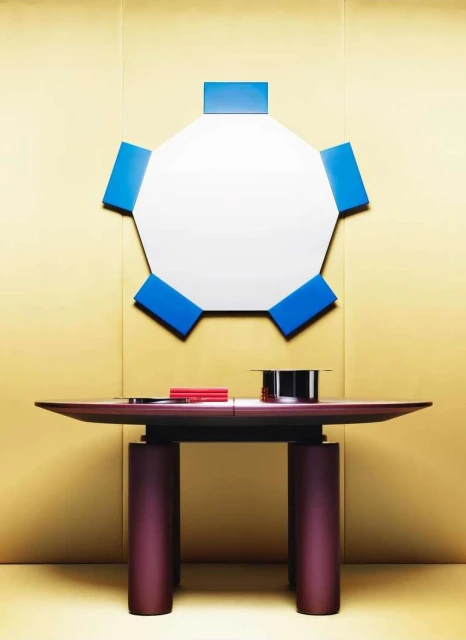 Contemporary interior featuring a table with cylindrical legs, a geometric mirror with blue accents, and a gold background.