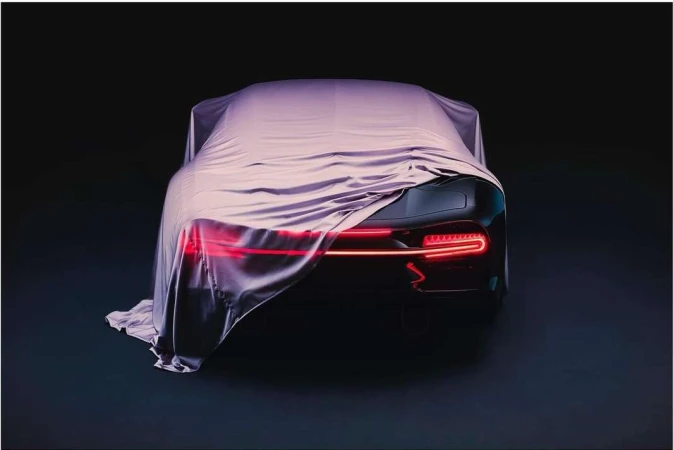 Rear view of a black sports car partially covered by a silver fabric sheet in a dark environment, showing sleek tail lights.