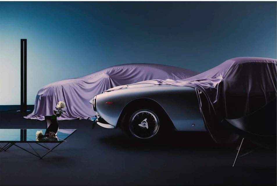 Two vintage cars, partially covered with lavender cloths, are staged in a dark studio against a two-tone gradient background.