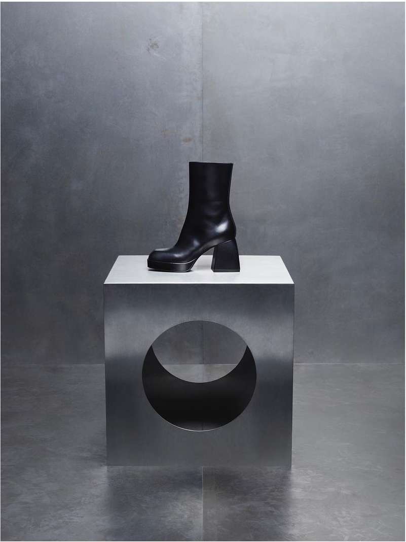 Platform Chelsea boot in black leather on a metallic cube with a circular cutout, set against a textured silver background.