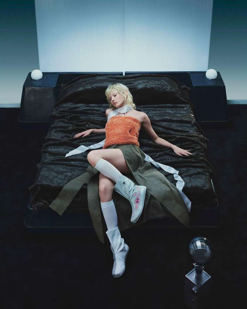 Woman in orange top and skirt lying on bed in futuristic room, wearing white boots and socks, lit by ambient and accent lighting.