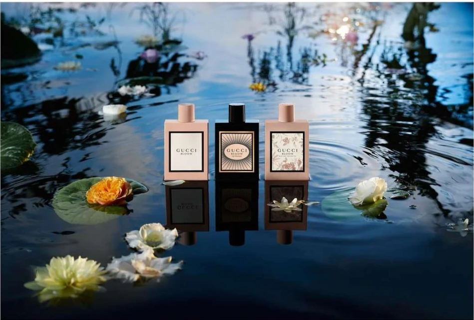 Three Gucci Bloom perfume bottles centered on dark, still water with flowers and lily pads under soft lighting. Beauty, product shot.