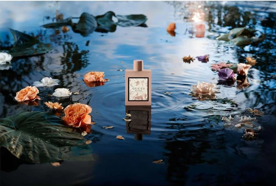 Gucci Bloom perfume bottle sits in tranquil water surrounded by vibrant flowers and lily pads, reflecting light with a serene aesthetic.