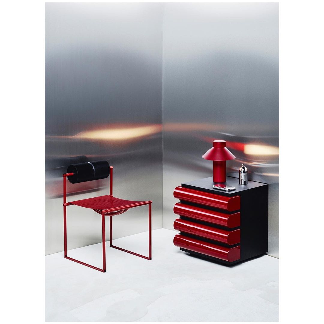 Minimalist interior featuring a red lamp on a black and red cabinet, alongside a red chair, set against a sleek silver wall.