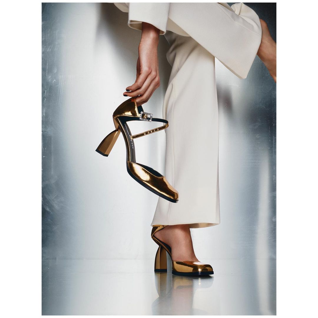 A fashionable woman wearing white pants is holding a shiny gold high-heeled shoe in her hand against a textured metallic background.