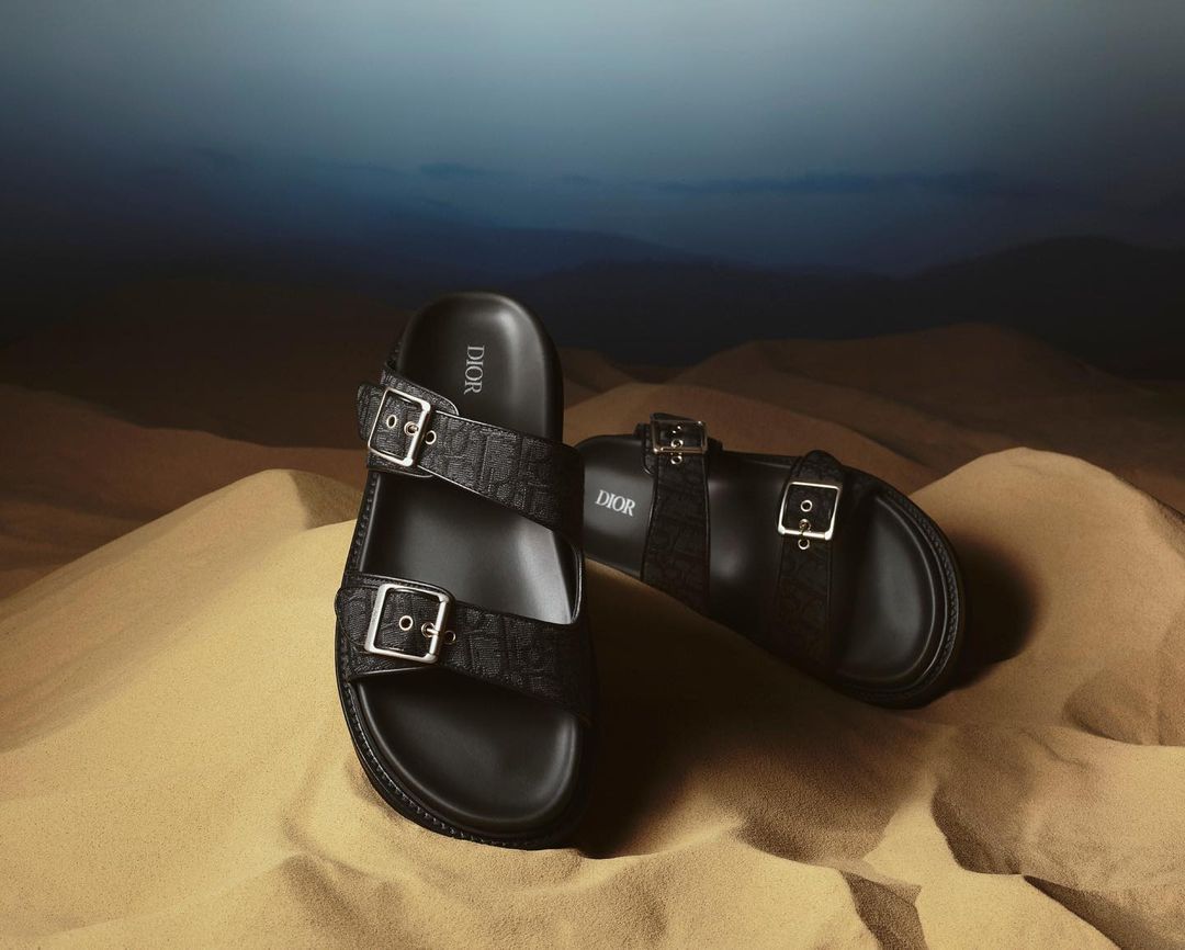 Close-up product shot of two black Dior slide sandals with silver buckles on a textured sand dune with a blue gradient sky backdrop.