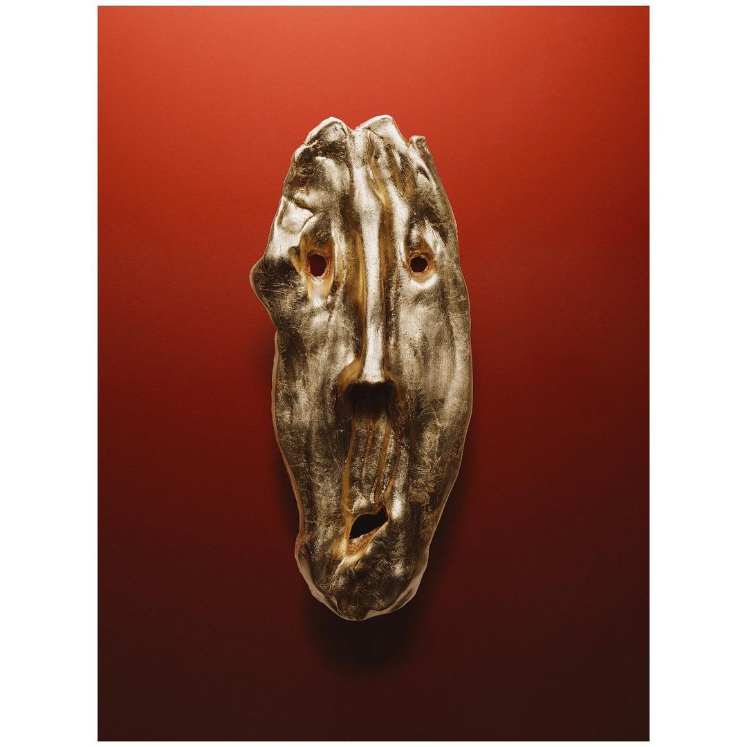 Striking gold-toned art mask with an expressive face on a rich red gradient background, creating a captivating contrast of form and color.