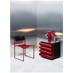 A red steel chair with black cylindrical headrest beside a matching red bedside table against brushed metal walls.